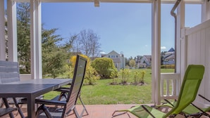 A covered patio with a table and chairs, a green lounge chair, and a view of a residential area with houses and greenery. - Comfort apartment with 1 bedroom and terrace on the first floor (Ostseebad Binz)