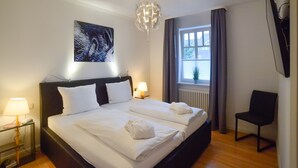 A hotel room with a large bed, bedside lamps, a ceiling light, a window with curtains, a chair, and a wall-mounted TV. - Comfort plus apartment with 1 bedroom and balcony on the upper floor (Ostseebad Binz)