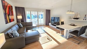 A modern living room with a sofa, TV, dining table, and a view of the outside. - Comfort plus apartment with 1 bedroom and balcony on the upper floor (Ostseebad Binz)