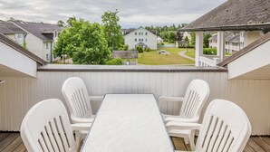 Een overdekt terras met een tafel en vier stoelen, met uitzicht op een woonwijk met huizen en een gazon.