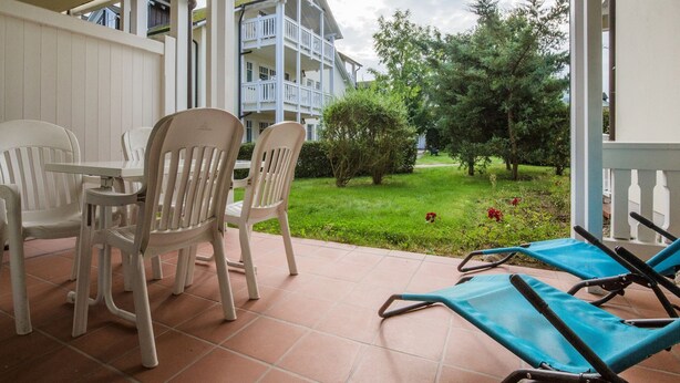 A patio with white plastic chairs and a blue lounge chair, a tiled floor, and a view of a building and greenery. - Comfort apartment with 1 bedroom and terrace on the first floor (Ostseebad Binz)