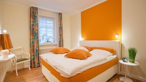 A neatly arranged bedroom with a large bed, a bedside table, a chair, and a window with curtains. - Comfortplus vacation apartment with 1 bedroom and terrace on the first floor (Ostseebad Binz)