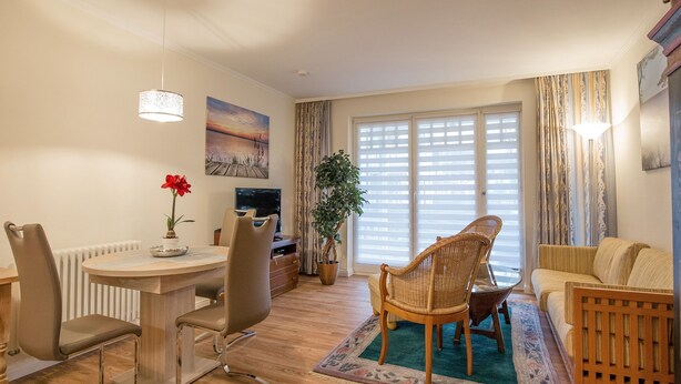 A modern living room with a dining area, a large window with blinds, a television, and a potted plant. - Comfortplus vacation apartment with 1 bedroom and terrace on the first floor (Ostseebad Binz)