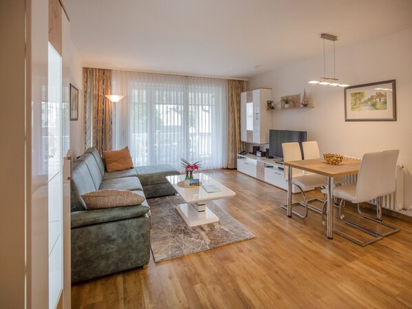 Un salon moderne avec un canapé d’angle gris, une table basse et un coin repas comprenant une table en bois et des chaises blanches.