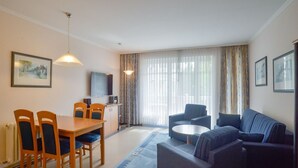 A modern living room with a dining area, a flat-screen TV, and a large window with curtains. - Comfort apartment with 1 bedroom and balcony on the upper floor (Ostseebad Binz)