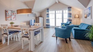 A modern living room with a blue sofa, a dining area with a wooden table, and a kitchen with a hanging light fixture.