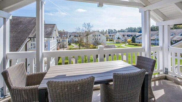 A covered patio with a table and wicker chairs, overlooking a residential area with houses and a well-maintained lawn.