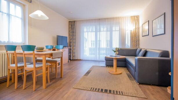 Uma sala de estar moderna com uma mesa de jantar de madeira, cadeiras azuis, um sofá cinzento e uma televisão.