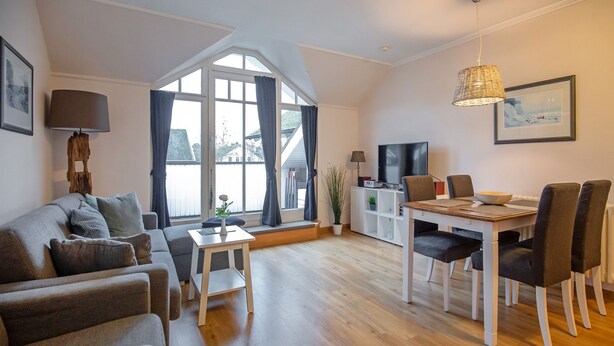 A modern living room with a sofa, dining table, and chairs. There is a TV, a lamp, and a plant. - Komfortplus Ferienwohnung mit 1 Schlafzimmer und Balkon im Dachgeschoss (Ostseebad Binz)