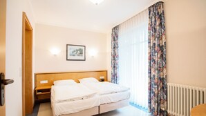 A hotel room with a double bed, a wooden headboard, a framed picture on the wall, a window with floral curtains, and a radiator.