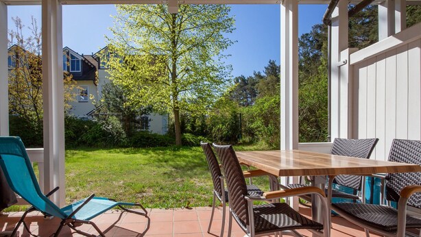 A covered patio with a wooden table and chairs, a blue lounge chair, and a view of a green lawn and trees.