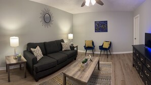 A living room with a dark sofa, two armchairs, a coffee table, a TV on a black cabinet, and a ceiling fan.