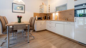 Eine moderne Küche mit weißen Schränken, einer Holzausziehtafel und einem Siemens-Kühlschrank. Ein Esstisch mit Stühlen und eine Blumenvase sind ebenfalls vorhanden.