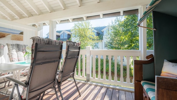 Eine überdachte Terrasse mit zwei Stühlen und einem Tisch, ein Holzdeck und ein weißer Zaun mit Blick auf eine Wohngegend.
