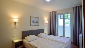 A hotel room with a bed, two bedside tables, a nightstand, a window with curtains, and a ceiling light. - Comfort apartment with 1 bedroom and terrace on the first floor (Ostseebad Binz)