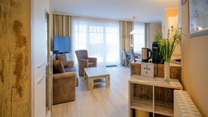 A modern living room with a sofa, chairs, a coffee table, and a TV. There is a wooden cabinet with a plant and a kettle on top. - Comfortplus vacation apartment with 1 bedroom and terrace on the first floor (Ostseebad Binz)