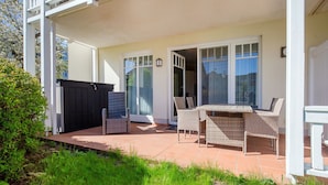 A covered patio with wicker furniture, a black gate, and a view of a building through glass doors. - Comfortplus vacation apartment with 1 bedroom and terrace on the first floor (Ostseebad Binz)