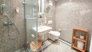 Un baño moderno con cabina de ducha de vidrio, un lavabo, un inodoro y un estante de madera con artículos de aseo.