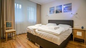 Een net opgemaakt bed met witte lakens, een houten nachtkastje met een klok, en een stoel met een kussen.