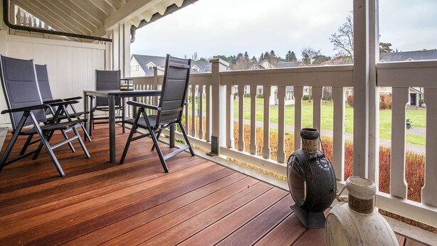 Een houten terras met buitenmeubilair, een witte balustrade en uitzicht op een woonwijk.