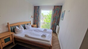 Ein Schlafzimmer mit einem hölzernen Bett, weißer Bettwäsche, einem Nachttisch, einem schwarzen Teppich und Blick auf Grün hinter dem Fenster.