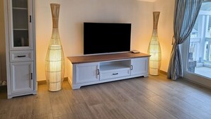Ein modernes Wohnzimmer mit einem Flachbildfernseher auf einem Holz-TV-Schrank, flankiert von hohen, zylindrischen Stehlampen.