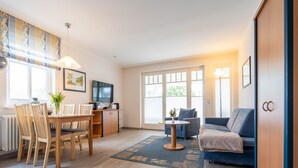 A modern living room with a dining area, a sofa, a TV, and a large window. - Comfort apartment with 1 bedroom and balcony on the upper floor (Ostseebad Binz)