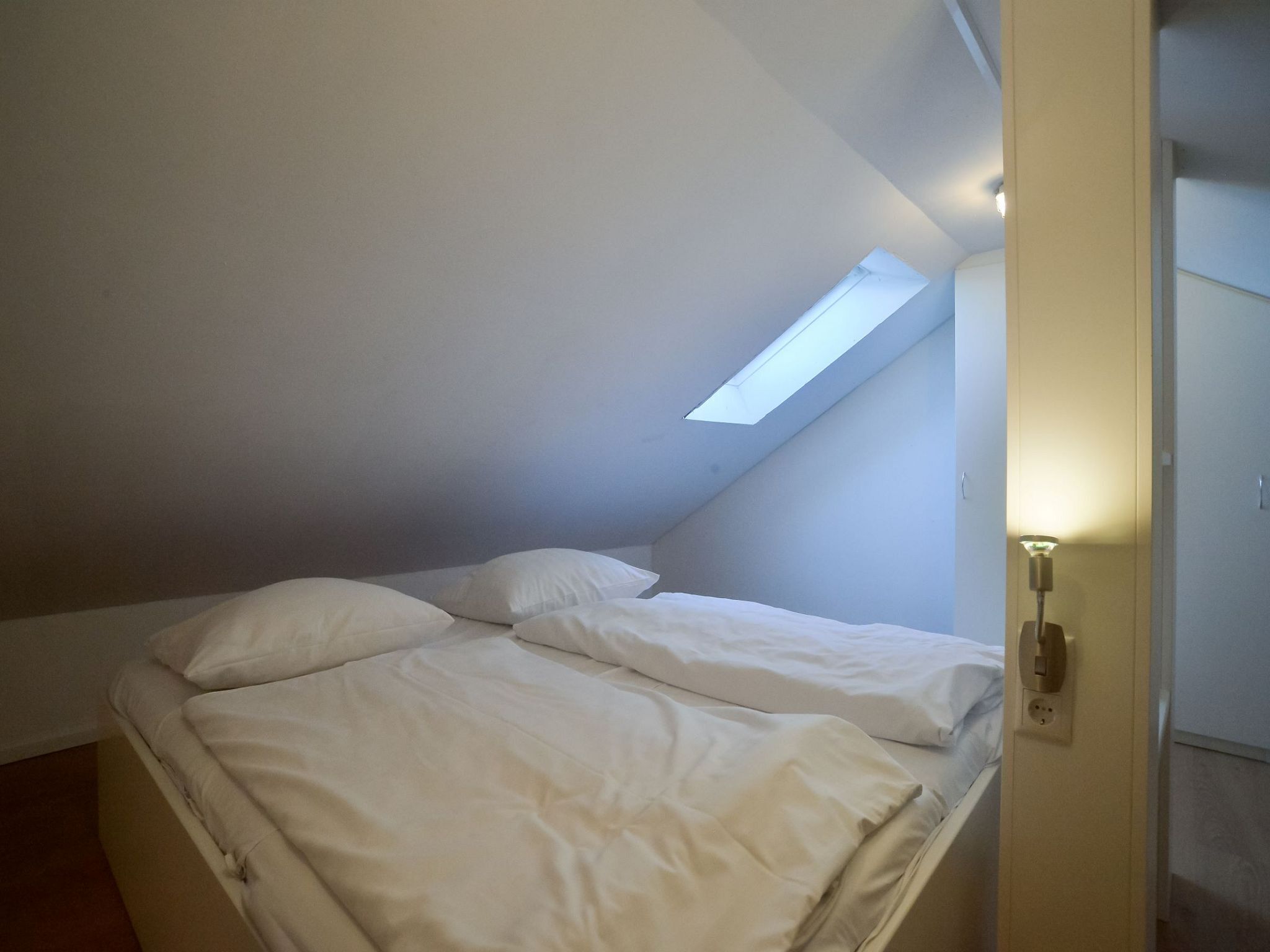 Une petite pièce faiblement éclairée, avec un lit simple, une lucarne et une lampe de chevet.