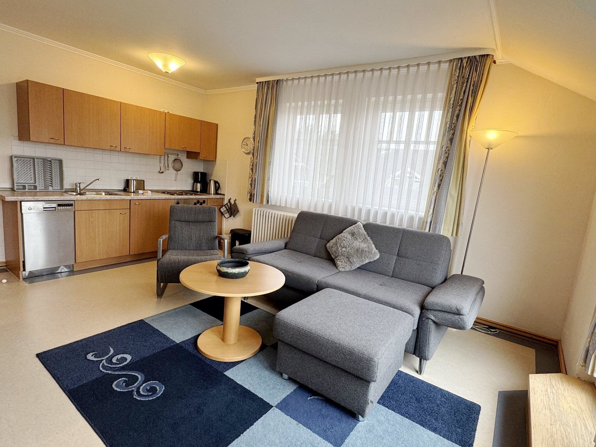 Un espace de vie compact comprenant un coin cuisine, un canapé gris, une table basse ronde et un tapis à motifs.