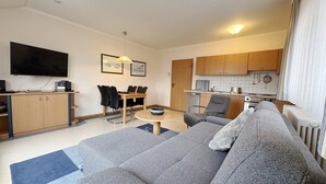 A modern living room with a grey sofa, a dining table with chairs, a kitchen area with wooden cabinets, and a flat-screen TV mounted on the wall. - Comfort apartment with 1 bedroom and balcony on the top floor (Ostseebad Binz)