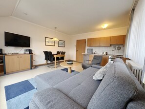 A modern living room with a grey sofa, a dining table with chairs, a kitchen area with wooden cabinets, and a flat-screen TV mounted on the wall. - Comfort apartment with 1 bedroom and balcony on the top floor (Ostseebad Binz)