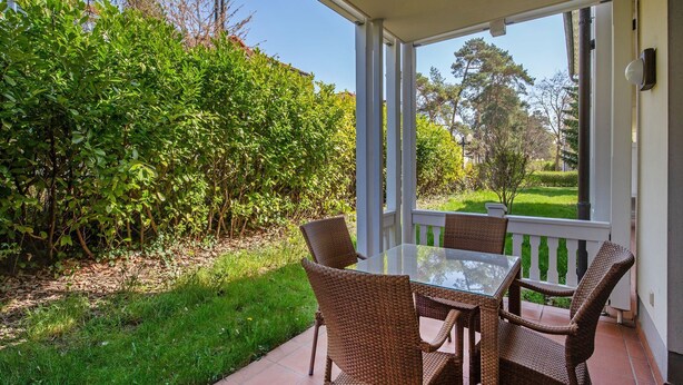 Une terrasse extérieure avec une table en verre et des chaises en osier, entourée d’un jardin et offrant une vue sur des arbres et un bâtiment.