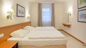 Une chambre d’hôtel avec deux lits, une tête de lit en bois, une table de chevet, une petite table, une lampe fixée au mur et une fenêtre avec des rideaux.