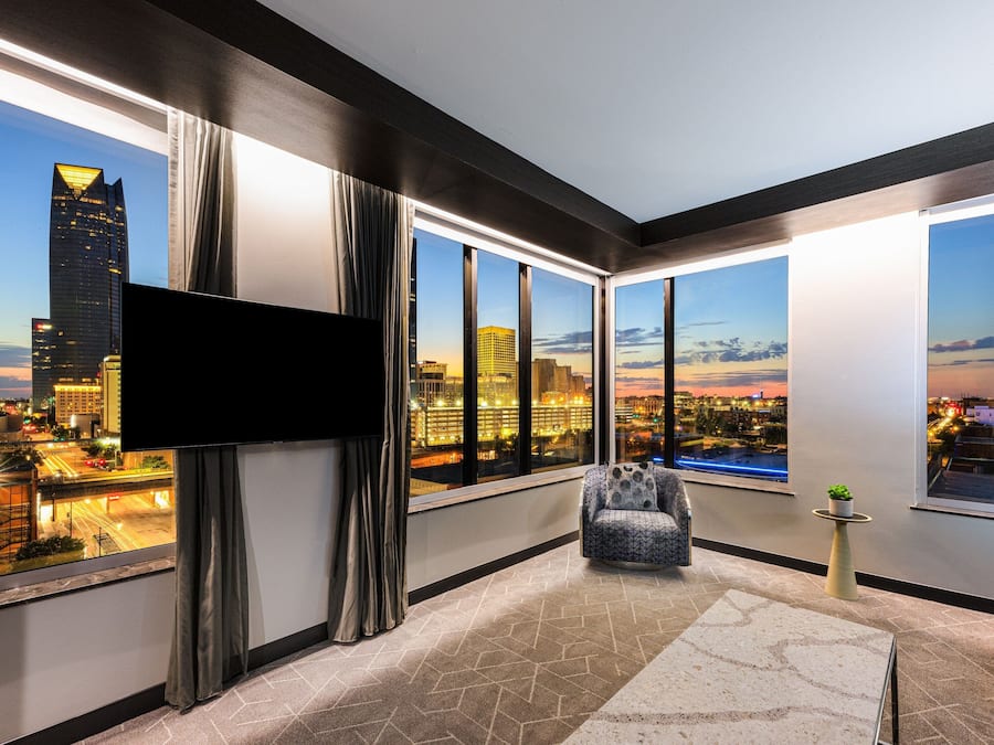 Habitación de hotel moderna con ventanas amplias, un televisor de pantalla plana, un sillón cómodo y una pequeña mesa con una planta.