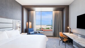 A hotel room with a large bed, a desk, a chair, a TV, and a view of the city through large windows. - Renaissance Oklahoma City Downtown Bricktown Hotel (Oklahoma City)
