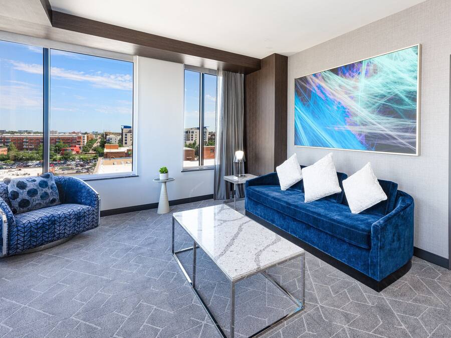 A modern hotel room with a large window, a blue sofa, a white coffee table, and a city view.