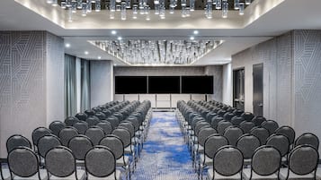 Een vergaderzaal met rijen stoelen, een groot scherm en een kamerbreed tapijt met een patroon.