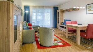 A modern living room with a sofa, armchair, and dining table. There is a window with curtains, a bookshelf, and a framed picture on the wall. - Comfort plus apartment with 1 bedroom and balcony on the upper floor (Ostseebad Binz)