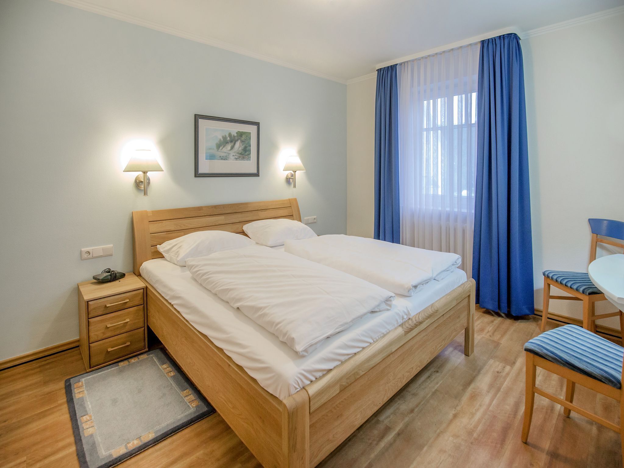 A neatly made bed with white linens, a wooden headboard, and two bedside lamps. There is a small bedside table with a lamp, a chair, and a rug on the floor.