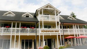 A hotel with a balcony, a sign reading "DÜNENPARK BINZ", and outdoor seating with red umbrellas. - Comfort plus apartment with 1 bedroom and balcony on the upper floor (Ostseebad Binz)