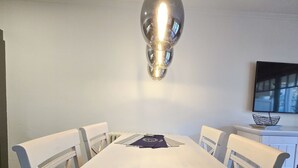Ein Essbereich mit einem langen Tisch, vier Stühlen, einer modernen Pendelleuchte, einem weißen Schrank mit einer Dekoration und einem Flachbildfernseher an der Wand.
