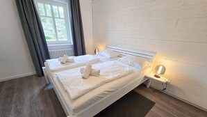 Ein Doppelbett mit weißen Bettwäsche und Handtüchern, ein Nachttisch mit Lampe sowie Blick auf Grün durch ein Fenster.