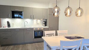 Eine moderne Küche mit grauen Schränken, einem Einbauofen, einer Mikrowelle und einem Spülbecken. Ein Esstisch mit weißen Stühlen und Pendelleuchten ist vorhanden.