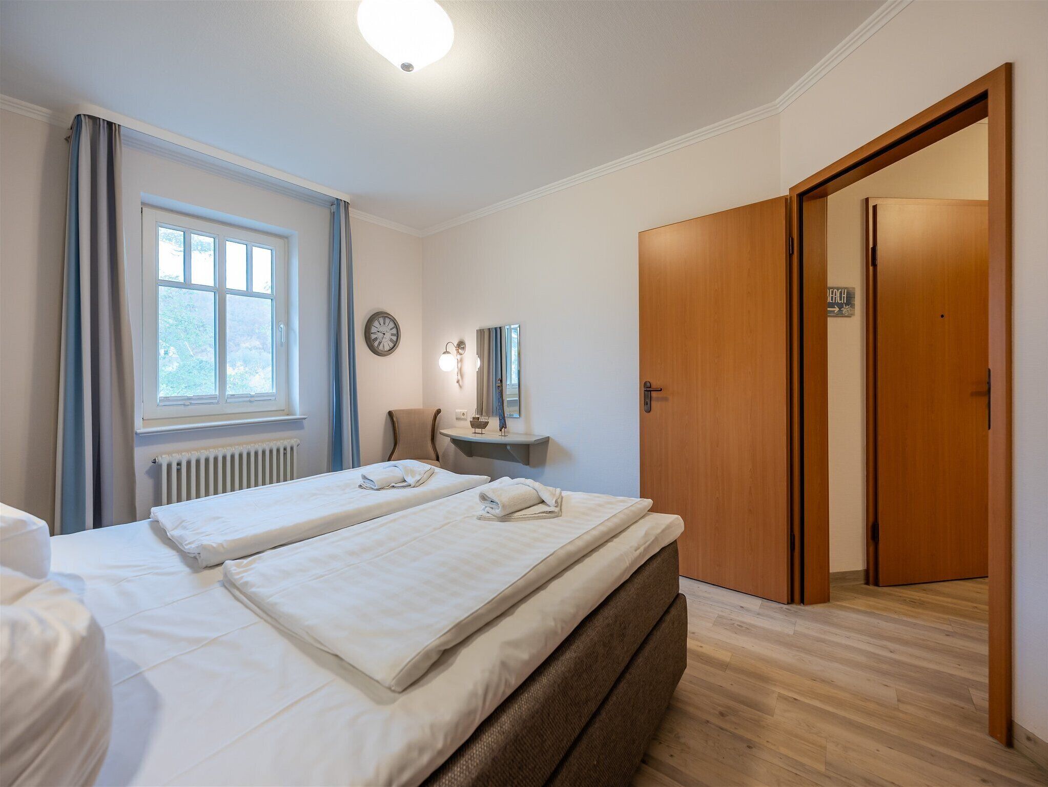 A hotel room with two beds, a desk, a chair, a clock, and a door.