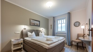Ein ordentlich eingerichtetes Schlafzimmer mit einem großen Bett, Nachttischen, einem Sessel und einem Fenster mit Vorhängen.