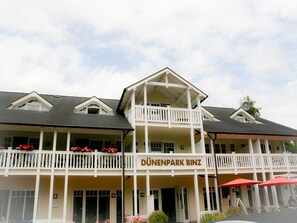 Ein Hotel mit Balkon, einem Schild mit der Aufschrift "DÜNENPARK BINZ" und Terrassenmöbeln mit roten Sonnenschirmen.