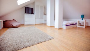 Une pièce moderne et bien éclairée, avec un plafond en pente, un téléviseur posé sur une commode blanche, un lit avec une lampe bleue et un grand tapis beige.
