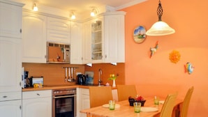 Een keuken met witte kastjes, een houten eettafel en oranje stoelen. Daarboven hangt een lamp.