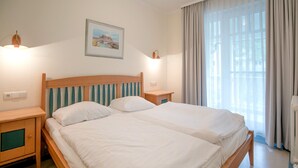 A double bed with wooden headboard and footboard, two bedside tables, a nightstand, a framed picture on the wall, and a window with curtains. - Comfort apartment with 2 bedrooms and balcony on the upper floor (Ostseebad Binz)