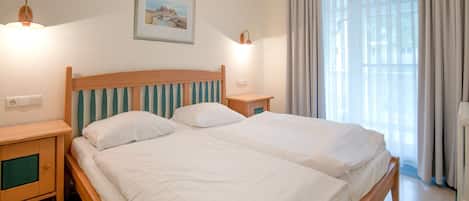 A double bed with wooden headboard and footboard, two bedside tables, a nightstand, a framed picture on the wall, and a window with curtains.